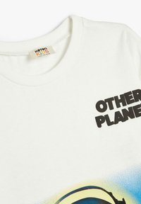 Valkoinen puuvillapaita lyhyillä hihoilla, jossa on suuri musta painatus sanoista "OTHER PLANET" ja värikäs graafinen kuva.