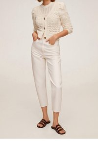 Jasnobeżowy sweter z dzianiny, zdobiony detalami i guzikami, w zestawie z off-white spodniami o wysokim stanie i brązowymi sandałami na paski.