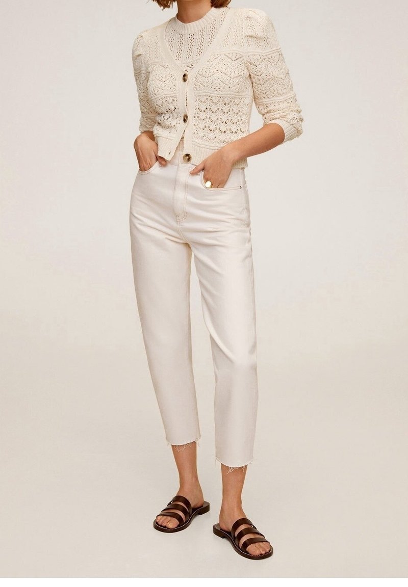 Jasnobeżowy sweter z dzianiny, zdobiony detalami i guzikami, w zestawie z off-white spodniami o wysokim stanie i brązowymi sandałami na paski.