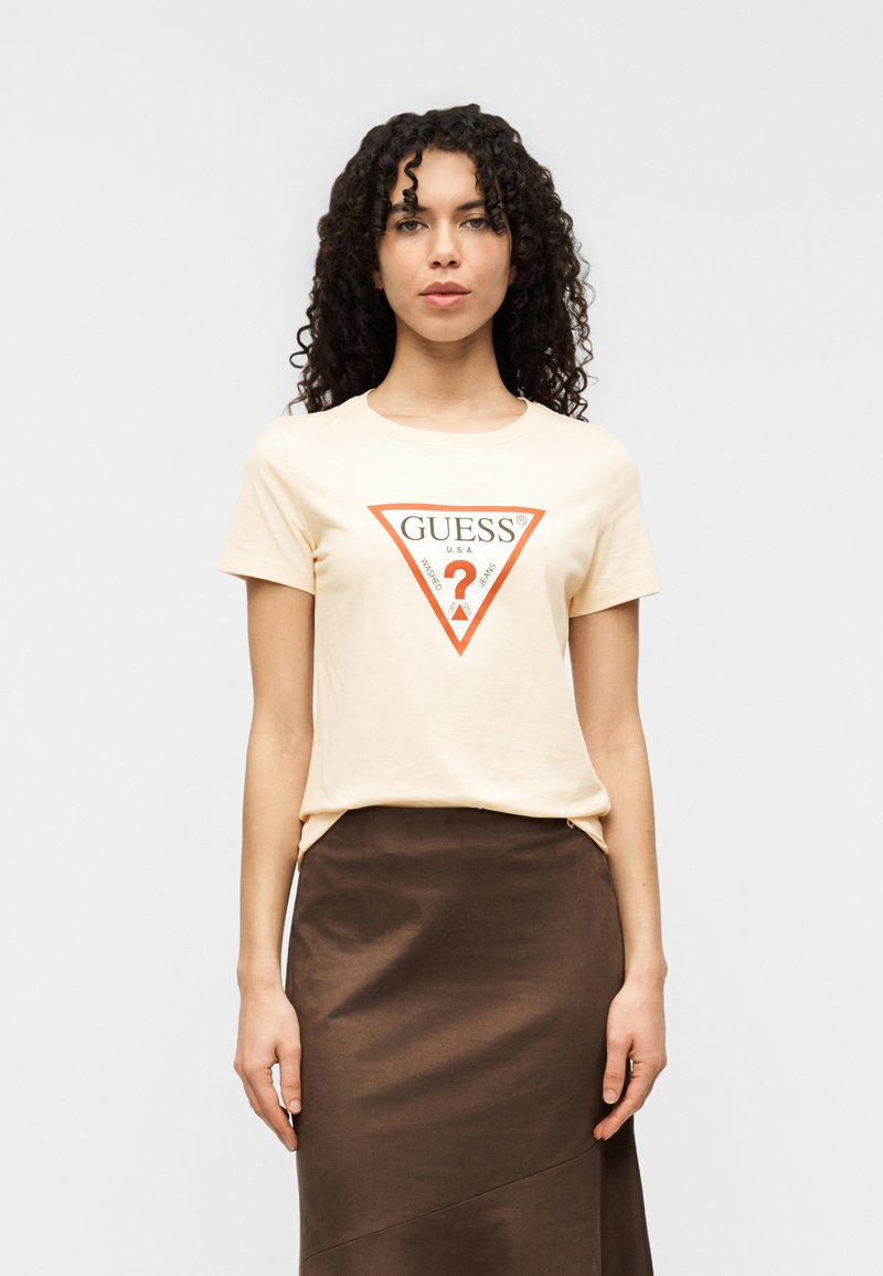 Μπεζ βαμβακερό T-shirt με κόκκινο τριγωνικό λογότυπο που περιλαμβάνει τη λέξη "GUESS" και ένα ερωτηματικό στο κέντρο. Συνδυασμένο με σκουρό καφέ φούστα.