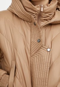 Beige pufferjack met een geborduurd patroon, voorzien van een hoge kraag, een ritssluiting en zilveren drukknoopjes. Zachte, gladde textuur.