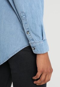 Ljusblå denimskjorta med lång ärm, med knäppbara manschetter och en något rundad nederkant; tillsammans med mörk tyg under.