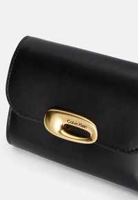 Juodas odinis piniginė su apvaliu auksiniu užsegimu. Yra siūtos kraštinės ir iškilmingas logotipas "Calvin Klein" ant užsegimo.
