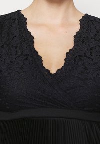 Czarna sukienka z koronkowym gorsetem i wycięciem w kształcie litery V oraz plisowanym dołem. Materiał ma strukturę z kwiatowym wzorem.