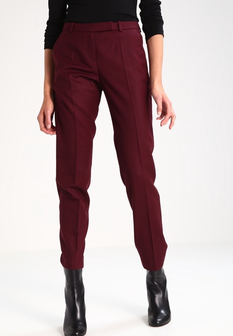 Femme portant un pantalon ajusté bordeaux avec pli frontal, un haut noir et des bottines noires, debout devant un fond uni.
