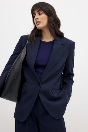 Marinblå blazer med skarpa slag, enkel knappstängning och två framfickor. I kombination med en mörk ribbad topp och en svart väska.