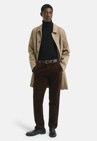 Vaaleanruskea trench-coat mustan poolopaitaan yhdistettynä ruskeisiin cordura-housuihin ja mustiin kenkiin. Malli seisoo kädet taskuissa.