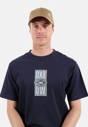Homme aux yeux bleus portant une casquette beige et un t-shirt bleu marine avec le texte blanc "OXB OW" et un dessin au centre, debout devant un fond blanc.