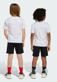 Dvě děti oblečené v bílých sportovních tričkách, černých šortkách a barevných ponožkách, stojící zády k sobě. Hladká textura látky, jednoduchý design.