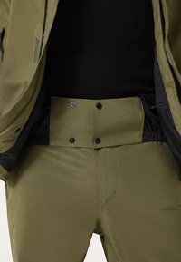 Oliwkowe spodnie outdoorowe o gładkiej fakturze, z zapięciami na napy i elastycznym pasem, połączone z czarną kurtką górną.