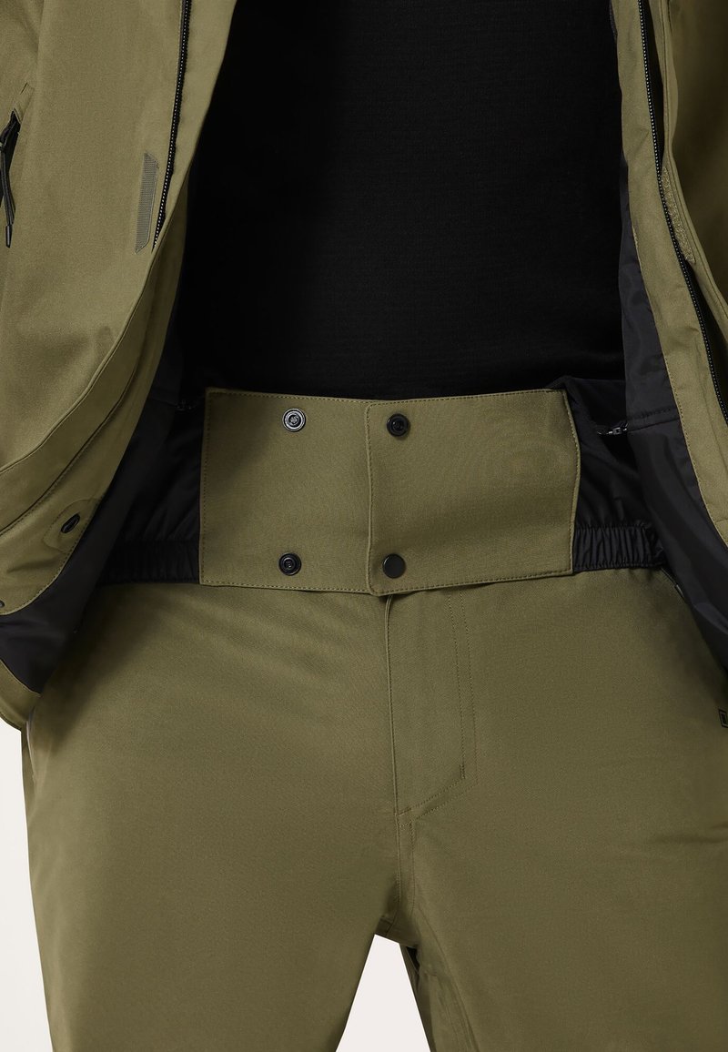 Olivgrüne Outdoor-Hosen mit glatter Textur, ausgestattet mit Druckknopfverschlüssen und elastischem Bund, kombiniert mit einer schwarzen Oberjacke.