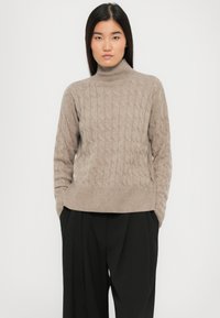 Frau trägt einen beigen, kabelgestrickten Rollkragenpullover und schwarze, gefaltete weitgeschnittene Hosen, mit den Händen in den Taschen, stehend vor einem weißen Hintergrund.