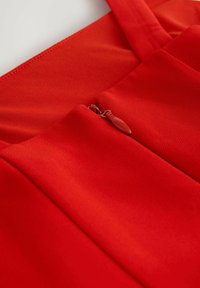 Rode stof met een gladde textuur, voorzien van een kleine ritssluiting en banden. Het ontwerp bevat plooien voor extra detail.