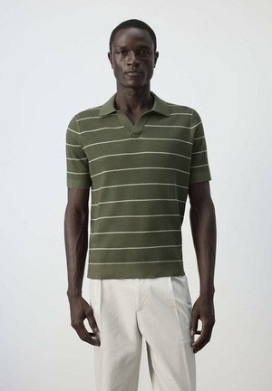Homme portant un polo vert à manches courtes avec des rayures blanches horizontales et un pantalon beige clair, debout devant un fond uni.