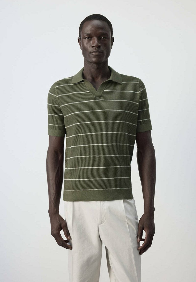 Homme portant un polo vert à manches courtes avec des rayures blanches horizontales et un pantalon beige clair, debout devant un fond uni.