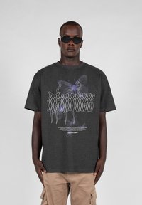 Camiseta gráfica de color gris oscuro con diseños de mariposas blancas y moradas y texto estilizado. Fabricada en tela suave, de manga corta y ajuste relajado.