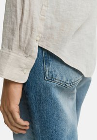 Camisa de lino beige claro con puños abotonados, combinada con pantalones vaqueros azules que tienen un bolsillo trasero y costuras en contraste.