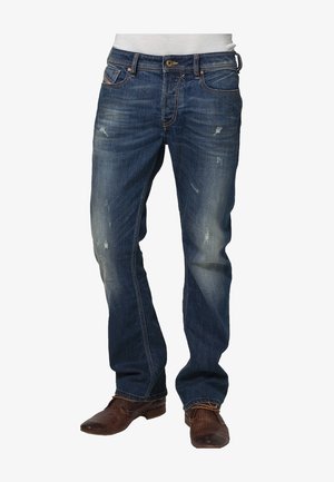 Jean bleu foncé délavé pour homme avec légère usure, porté avec des chaussures en cuir marron texturé et une chemise blanche rentrée.