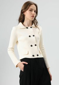 Veste de couleur crème avec un col rond, des boutons noirs, deux poches avant et une coupe cintrée. Le matériau présente une texture apparente.