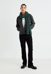 Vuxen manlig modell i en svart jeansjacka, vit t-shirt, svarta byxor och en grön texturerad scarf. Svarta skor fulländar outfiten.