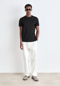 Camiseta de algodón negra con cuello redondo, combinada con pantalones blancos holgados. Modelo luciendo gafas de sol oscuras y zapatillas grises. Diseño simple.