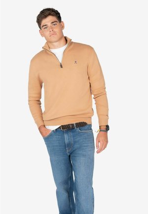 Suéter de punto beige con cuello de cremallera, textura suave. Combinado con jeans de mezclilla azules, que cuentan con un cinturón trenzado y un sutil detalle de reloj.