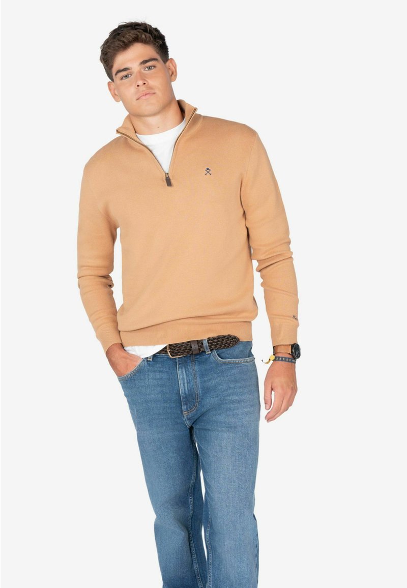Suéter de punto beige con cuello de cremallera, textura suave. Combinado con jeans de mezclilla azules, que cuentan con un cinturón trenzado y un sutil detalle de reloj.