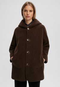Cappotto in pile marrone con cappuccio, dotato di grandi tasche frontali, chiusure a bottoni e un tessuto testurizzato. Silhouette semplice e rilassata.