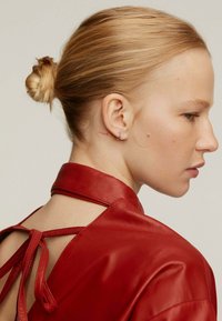 Prenda de cuero rojo con un alto cuello y espalda descubierta, con un cierre de lazo. Textura suave y mínimo detalle. Tatuaje sutil visible.