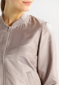 Chaqueta bomber de color taupe claro con un acabado de satén liso, que presenta un cierre de cremallera, un cuello de canalé y bolsillos laterales.