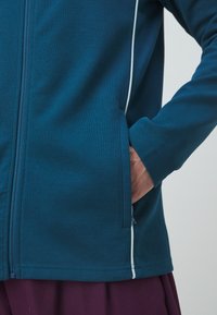 Mėlynas užtrauktuku užsisegamas džemperis su lygiu paviršiumi, puikuojantis baltomis siūlėmis ir šonine kišene, integruota į dizainą.