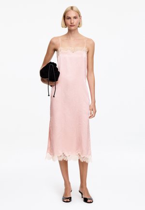 Mujer con un vestido de tirantes rosa pálido con encaje, sosteniendo una bolsa negra con cordón, y calzando tacones abiertos negros.