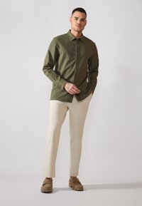 Camisa de botones verde oliva en una tela texturizada, combinada con pantalones crema de corte tapered y zapatos marrones de deslizar. El modelo está de pie contra un fondo claro.