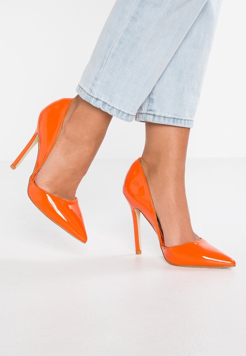 Pieds portant des chaussures à talons hauts pointues et brillantes de couleur orange, associées à un jean court bleu clair, sur un sol blanc.