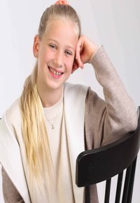 Leicht strukturierter beiger Pullover, der unter einem cremefarbenen Oberbekleidungsstück getragen wird und einen runden Halsausschnitt hat. Lange blonde Haare, entspannt neben einem schwarzen Stuhl sitzend.