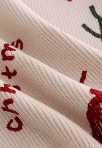 Zbliżenie jasnoróżowej materiału prążkowanego z czerwonymi i czarnymi haftowanymi literami oraz kształtami, ukazujące teksturę i fałdy.