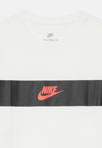 Bílý bavlněný tričko s horizontálním černým pruhem a červeným logem Nike uprostřed. Žebrovaný kulatý výstřih, hladká textura.