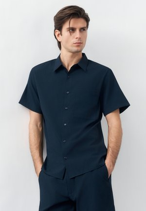 Mann mit dunklen Haaren, der ein marineblaues, kurzärmeliges Hemd mit Knöpfen und passende Hosen trägt, steht mit den Händen in den Taschen und blickt seitlich.