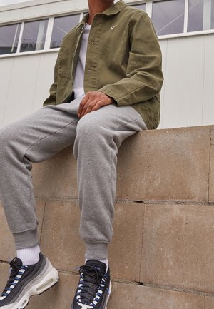 Mężczyzna w oliwkowo-zielonej kurtce, białej koszulce, szarych spodniach dresowych i czarno-białych trampkach siedzący na betonowym murku na zewnątrz.