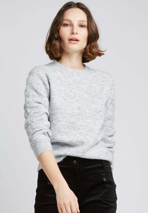 Pull en tricot gris clair avec un col rond, manches texturées et poignets côtelés, associé à un pantalon en velours côtelé noir.