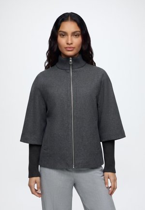 Femme aux longs cheveux foncés porte une veste zippée gris foncé à larges manches courtes par-dessus des manches longues côtelées noires moulantes et un pantalon gris clair.
