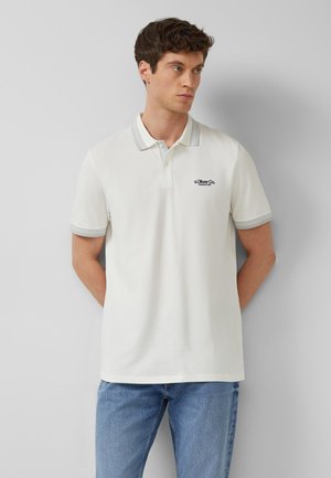 Mężczyzna noszący białą koszulkę polo z krótkim rękawem, szarym kołnierzykiem i mankietami oraz jasnoniebieskie dżinsy, stojący z rękami z tyłu.