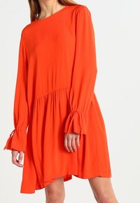 Oranssi pitkähihaine mekko, jossa on kerätty vyötärö, levenevä helma ja nauhat hihansuissa. Valmistettu kevyestä kankaasta, jossa on sileä tekstuuri.