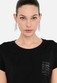 Kvinde med heterokromi, iført funkende, hængende øreringe og en sort top med et lille rhinestonesdesign på brystet.