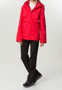 Veste imperméable rouge avec capuche, fermeture éclair et manches longues, associée à un pantalon noir et des chaussures de randonnée noires.
