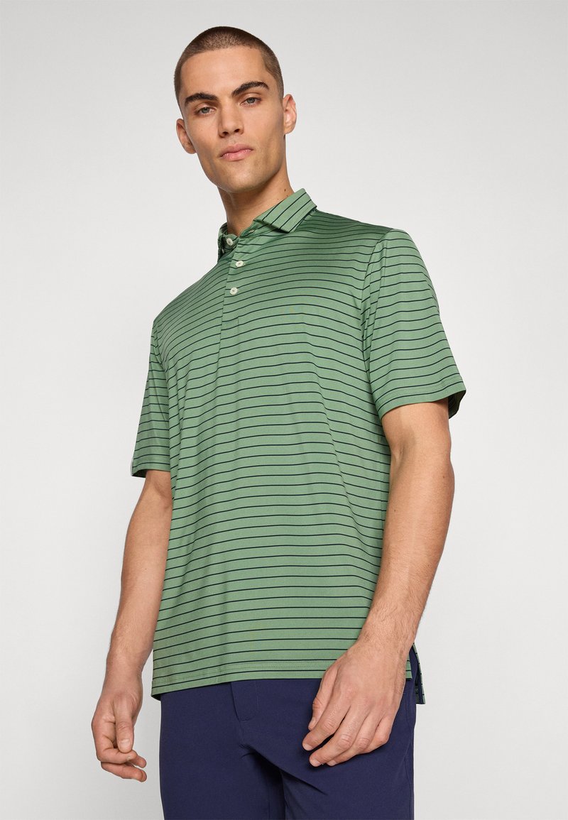 Man med kortärmad polo i grön och svart randig stil och marinblå byxor, stående mot en enkel ljus bakgrund.