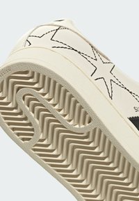 Baskets en cuir de couleur crème avec des motifs d'étoiles noires. La semelle en caoutchouc texturée présente un design à chevrons pour une meilleure adhérence.