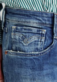 Blå denimjeans med en distinkt bakficka med en V-formad design. Tyget har en lätt blekt textur och synliga sömmar.