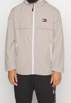 Beige capuchonjas met witte ritssluiting en zakken, met een Tommy Jeans-logo op de borst, gedragen met een zwarte broek.