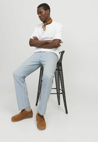 Hvit kortermet t-skjorte, lyseblå jeans, brune semskede sko og et brunt skjerf. Personen sitter på en svart perforert barstol.